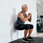 A senior man doing wall squat exercise for flexibility and strength, in a modern home gym setting, promoting stretching and daily fitness routines for seniors.