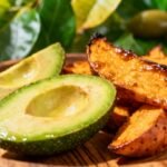 Ripe avocado slices and roasted sweet potatoes on wooden plate with green leaves and olives in background.